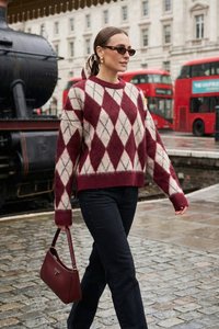 Kvinde iført en burgundy og beige argyle sweater og sorte bukser, bærende en burgundy håndtaske, går på en gade med røde dobbeltdækkerbusser.