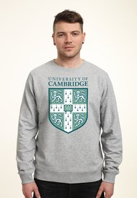 Felpa grigia con il blasone dell'Università di Cambridge. Il design include elementi color turchese e bianco con motivi di leone e freccia.