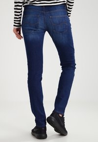 Slim-fit tmavě modré džíny s kontrastním prošitím a dvěma zadními kapsami, nošeny s černými teniskami a pruhovaným topem.