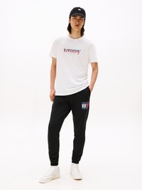 T-shirt blanc avec un logo "tommy" rouge et bleu, associé à un pantalon de jogging noir avec un accent de logo, et des baskets blanches. Tenue décontractée.