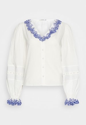 Blouse blanche à manches longues avec manches bouffantes, bordure en dentelle florale bleue sur le col en V et les poignets, devant boutonné, et détails discrets de couture horizontale sur les manches.