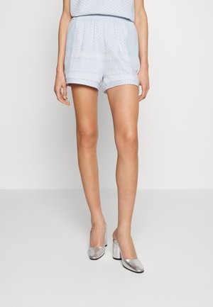 Femme portant un short bleu clair à motifs et des chaussures à talons blocs argentées, debout devant un fond clair uni.