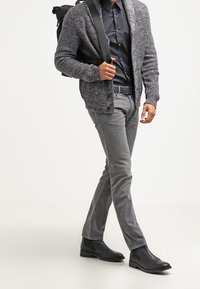 Grauer strukturierter Cardigan über einem schwarzen Hemd, graue Slim-Fit-Hose und schwarze Chelsea-Stiefel. Das Model hält mit einer Hand einen schwarzen Rucksack.