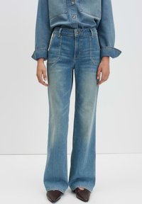 Jean en denim bleu clair taille haute, à jambes larges, avec des poches avant et une texture légèrement décolorée. Associé à une chemise en denim assortie.