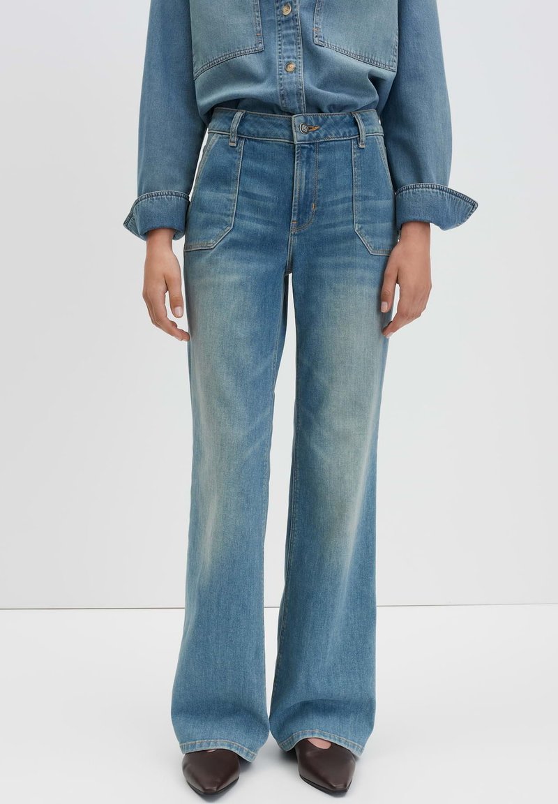 Jean en denim bleu clair taille haute, à jambes larges, avec des poches avant et une texture légèrement décolorée. Associé à une chemise en denim assortie.