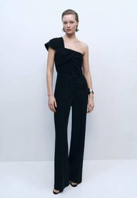 Black one-shoulder jumpsuit with bow detail, long wide legs, and structured soft fabric. Model wears a wristwatch and heeled sandals.
