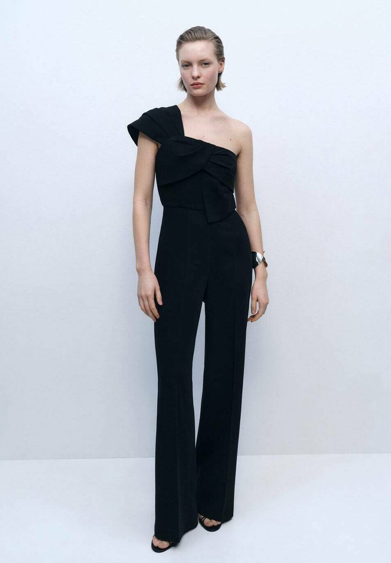 Black one-shoulder jumpsuit with bow detail, long wide legs, and structured soft fabric. Model wears a wristwatch and heeled sandals.