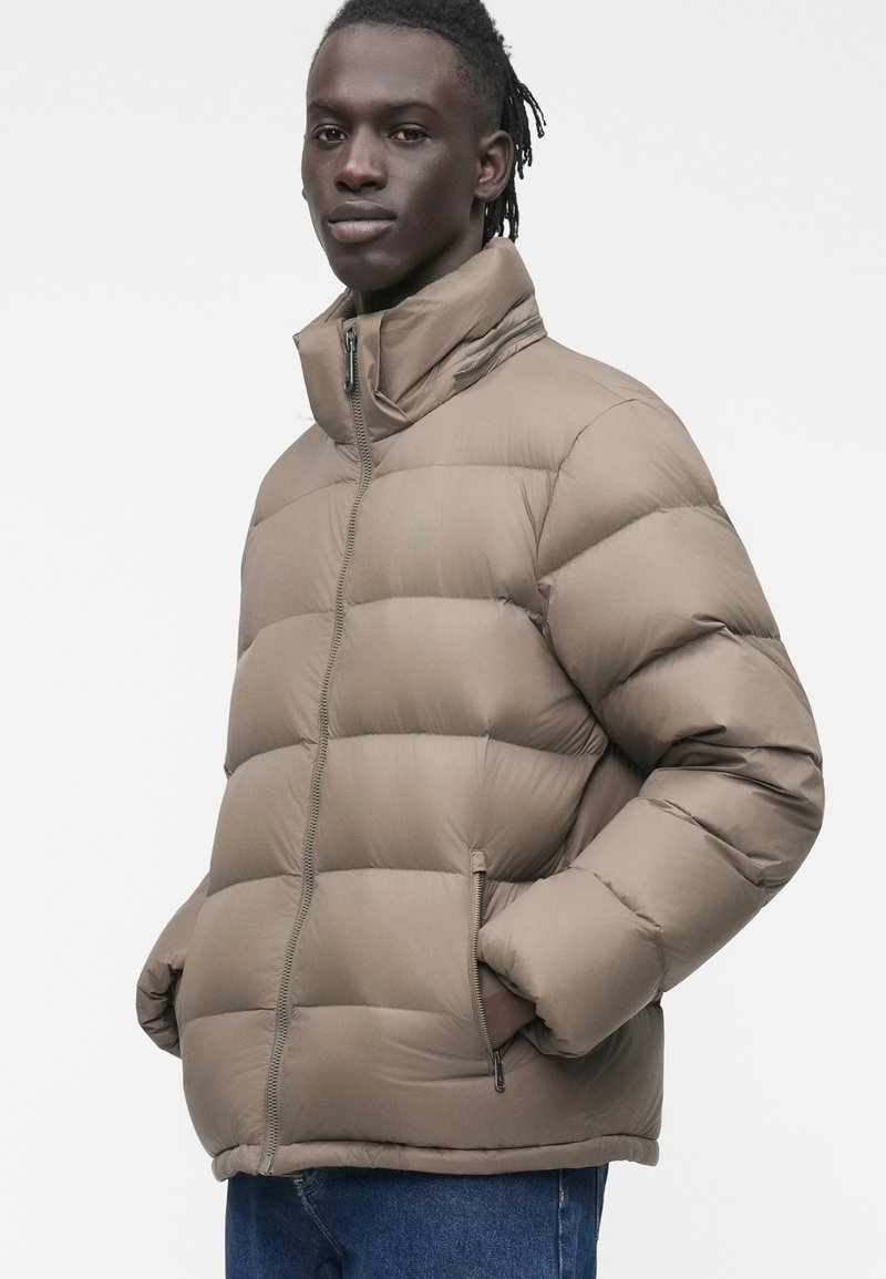 Junger Mann mit dunkler Haut und geflochtenem Haar trägt eine taupefarbene Steppjacke mit hohem Kragen und blaue Jeans, die Hände in den Jackentaschen.