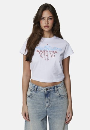 Młoda kobieta w białym krótkim T-shircie z napisem "Good For Nothing" i niebieską grafiką, w połączeniu z jasnoniebieskimi, wysokim stanem jeansami.