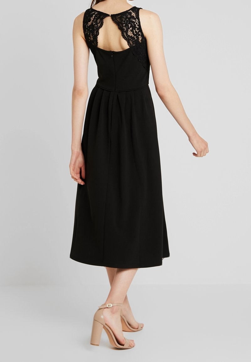 Femme portant une robe midi noire sans manches avec un détail en dentelle au dos et des sandales nude à talons hauts, debout devant un fond uni.