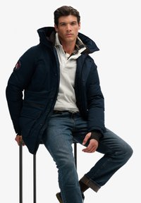 Doudoune bleu marine avec capuche, dotée de poches poitrine et latérales, portée sur un polo gris clair. Le modèle porte un jean bleu et des chaussures foncées.