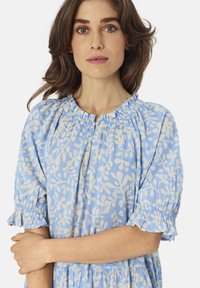 Světle modré šaty vyrobené z lehkého materiálu s bílým vzorem listů, s elastickým nařaseným výstřihem a volánovými rukávy.