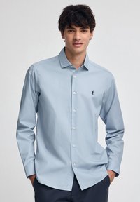 Giovane uomo che indossa una camicia azzurro chiaro con un piccolo logo scuro sul petto, in piedi con le mani nelle tasche dei pantaloni blu navy.