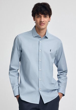 Junger Mann in einem hellblauen, kurzärmligen Hemd mit einem kleinen dunklen Logo auf der Brust, der mit den Händen in den Taschen seiner marineblauen Hose steht.