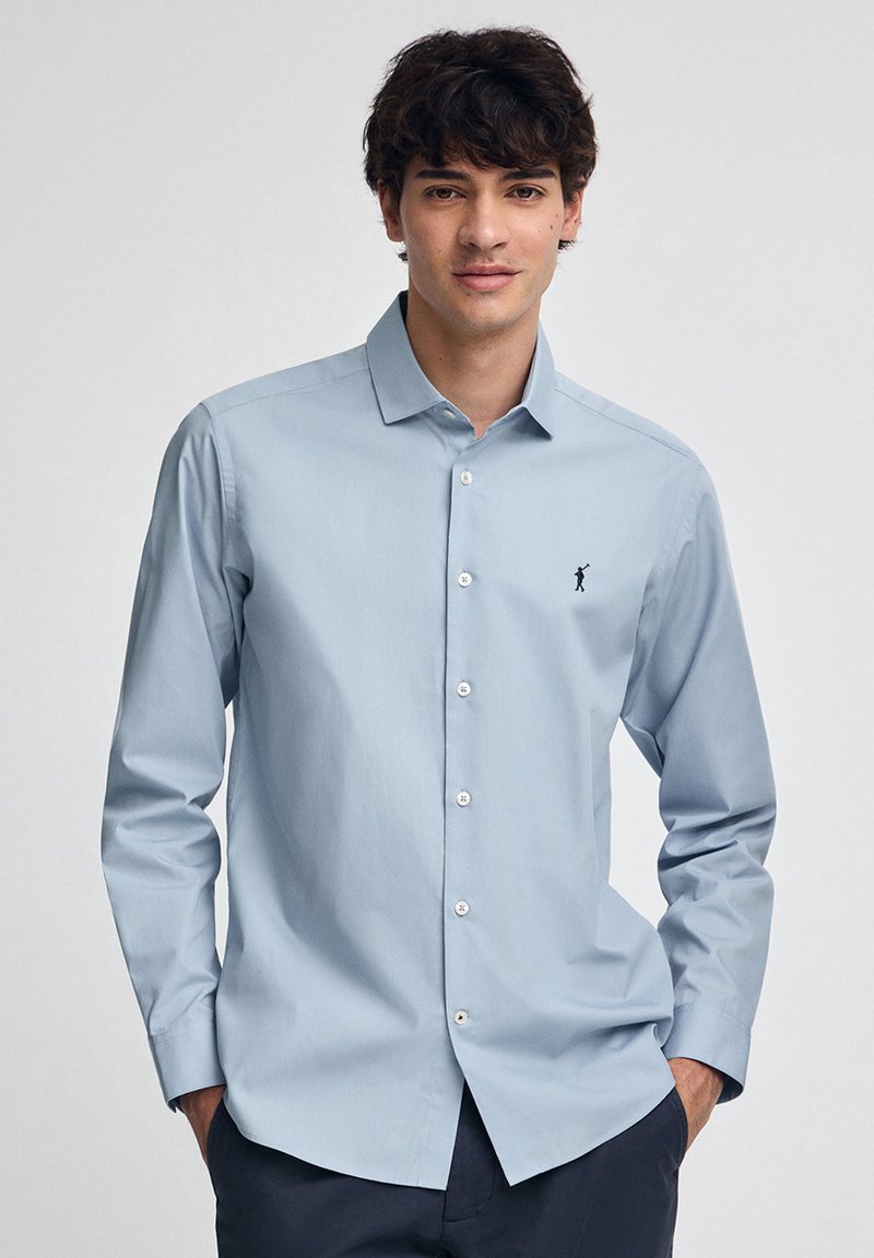 Giovane uomo che indossa una camicia azzurro chiaro con un piccolo logo scuro sul petto, in piedi con le mani nelle tasche dei pantaloni blu navy.