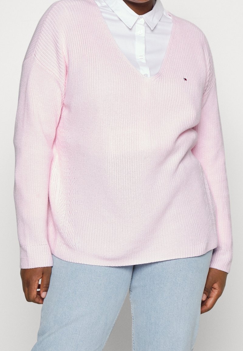 Personne portant un pull tricoté rose clair à col en V par-dessus une chemise blanche à col, et un jean bleu clair, debout devant un fond uni.