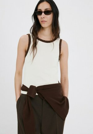 Donna con top bianco senza maniche con bordatura marrone, maglione marrone annodato in vita, pantaloni scuri e occhiali da sole rettangolari neri.