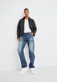 Svart kort jakke, hvit t-skjorte, falmede lys blå jeans og hvite sneakers, poserer mot en enkel hvit bakgrunn.