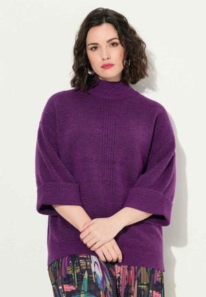Pull en maille violet avec un col montant et des manches courtes retournées. Il présente une texture côtelée et une coupe ample, associé à une jupe à motifs.