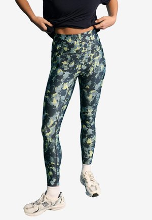 Person iført camouflage mønstrede leggings med høj talje, hvide sokker, hvide sneakers og løfter en sort skjorte for at afsløre taljen.