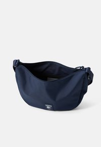 Mørkeblå Lacoste crossover-veske med glidelåsåpning og justerbar stropp, viser logo-patch foran mot hvit bakgrunn.