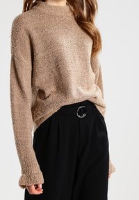 Pull en maille beige avec finition texturée, col montant et poignets côtelés, associé à un pantalon noir doté d'une boucle circulaire argentée.