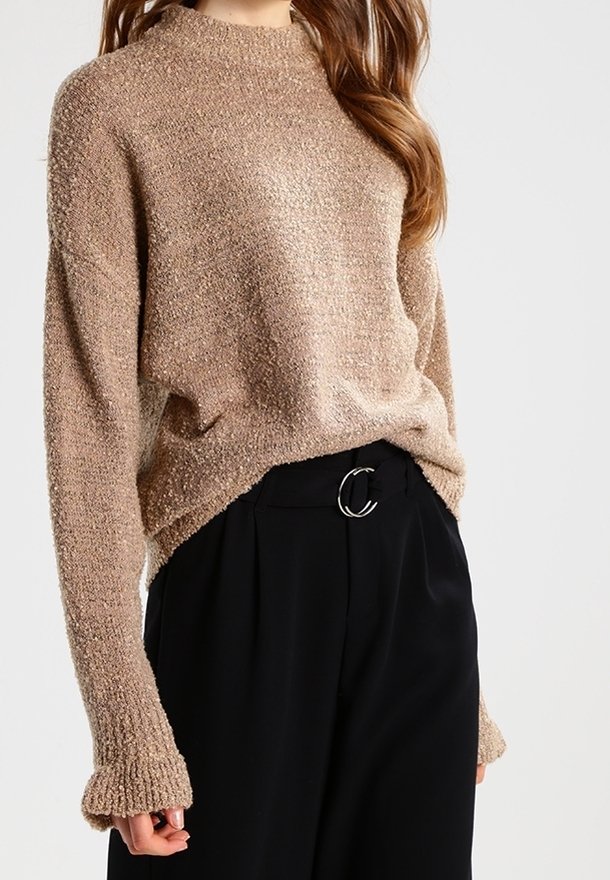 Pull en maille beige avec finition texturée, col montant et poignets côtelés, associé à un pantalon noir doté d'une boucle circulaire argentée.