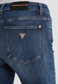 Jeans de mezclilla en azul oscuro con un acabado texturizado. Presenta un parche de logo metálico triangular y costuras visibles en los bolsillos.