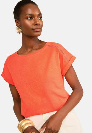 Žena oblečená v jasne oranžovom tričku s krátkym rukávom, veľké zlaté náušnice v tvare slnka a viaceré zlaté náramky, pózujúca s rukami pri páse.