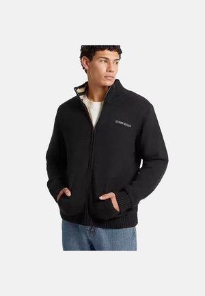 Junger Mann trägt einen schwarzen Reißverschluss-Pullover mit "Guess Jeans"-Logo, Hände in den Taschen, schaut leicht seitlich, vor einem einfarbigen Hintergrund.