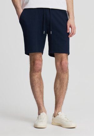 Uomo che indossa pantaloncini blu navy con coulisse, scarpe da ginnastica bianche e una maglietta bianca con le mani nelle tasche, in piedi contro uno sfondo chiaro e semplice.