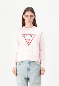 Sweatshirt rose avec un col rond et des manches longues. Il présente un logo central en triangle rouge avec le texte "GUESS" et un point d'interrogation à l'intérieur.