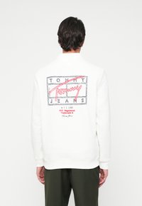 Mand iført hvid sweatshirt med Tommy Jeans-logo og rød tekst på ryggen, parret med mørkegrønne bukser, stående foran en ensfarvet baggrund.