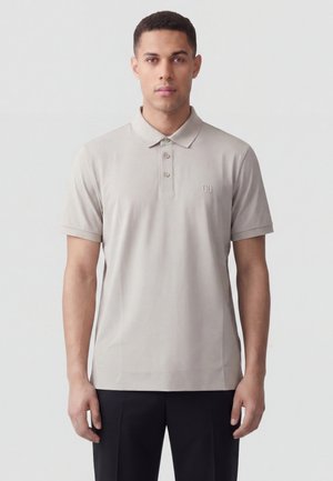 Giovane uomo che guarda avanti, indossa una polo a maniche corte grigio chiaro e pantaloni neri su uno sfondo chiaro uniforme.