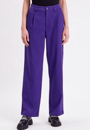 Femme portant un pantalon violet taille haute à jambes larges associé à des mocassins noirs épais avec des chaînes argentées, debout sur un fond blanc.