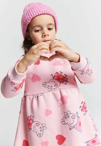 Junges Mädchen mit pinkem Hut und einem Kleid mit Herzmustern von Peppa Wutz, das mit ihren Händen eine Herzform bildet.