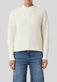 Eine Person trägt einen strukturierten, cremeweißen Rundhalsausschnitt-Pullover über einem weißen Hemd mit Kragen und mitteldunkelblauen, geraden Jeans.