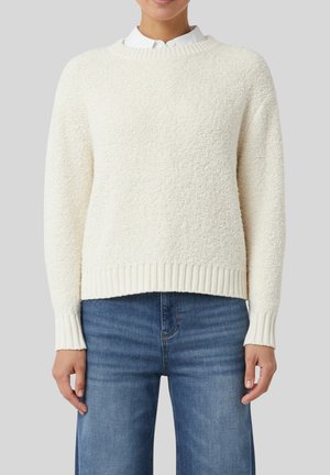 Osoba ubrana w teksturowany sweter w kolorze off-white z okrągłym dekoltem na białej koszuli z kołnierzykiem, do tego niebieskie dżinsy o prostym kroju.