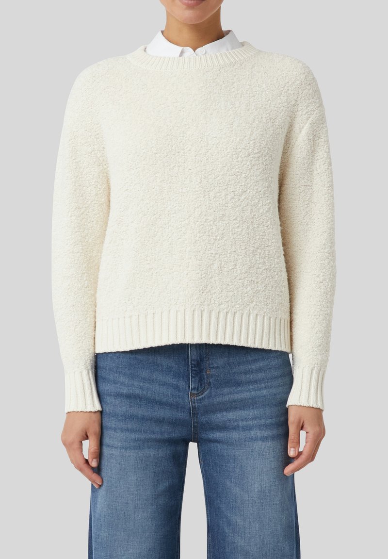 Eine Person trägt einen strukturierten, cremeweißen Rundhalsausschnitt-Pullover über einem weißen Hemd mit Kragen und mitteldunkelblauen, geraden Jeans.