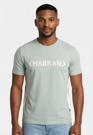 Hombre con cabello corto y barba, lleva una camiseta verde claro con el texto "CHABRAND" y jeans azul claro, de pie frente a un fondo blanco.