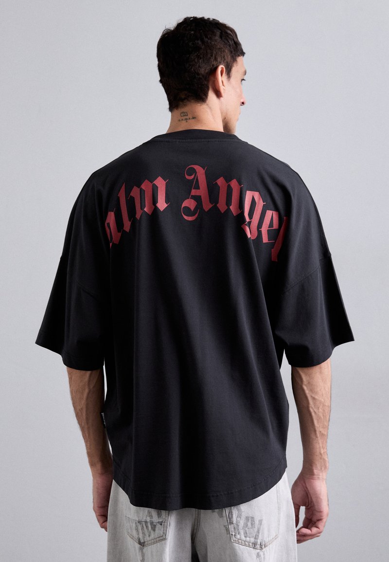 Must have lõtv T-särk lühikeste varrukatega, mille seljal on punase gooti fondiga tekst "Palm Angel". Kude näib sujuv ja pehme.