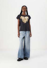 Korte mouwen zwarte T-shirt met gouden verenprint bij de halslijn, gecombineerd met wijde lichtblauwe jeans en zwarte chunky schoenen.