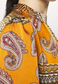 Blouse jaune à motifs avec des dessins paisley, présentant un tissu texturé, un col détaillé et un bord contrastant en rouge, noir et bleu.