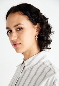 Un modèle aux cheveux bouclés porte une chemise rayée dans des tons clairs. Elle a des boucles d'oreilles en or et un maquillage naturel, mettant en valeur des taches de rousseur et une expression neutre.