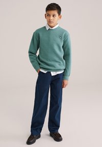 Zelený sveter s hexagonálnym vzorom, pod ním biela košeľa s golierom, kombinované s tmavomodrými nohavicami a tmavohnedými topánkami.