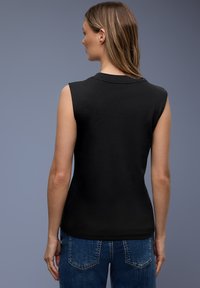 Femme aux cheveux raides châtain clair, portant un haut noir sans manches et un jean bleu, debout de dos sur un fond gris.