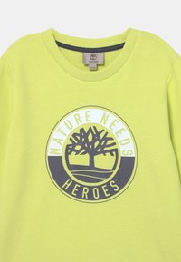 Sudadera amarilla brillante con un logo redondo que presenta un árbol y el texto "LA NATURALEZA NECESITA HÉROES". Cuello acanalado y tejido texturizado.