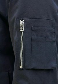 Černá látková kapsa se stříbrným zipem na straně, nad níž se nachází obdélníková klopová kapsa. Hladký povrch a čisté linie.