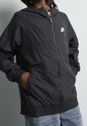 Person trägt eine schwarze Nike-Zip-up-Jacke, die Hände in den Taschen und hellblaue Jeans, steht vor einem schlichten hellgrauen Hintergrund.