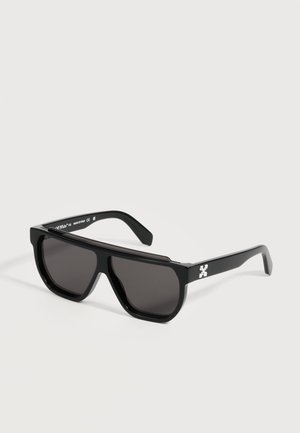 Lunettes de soleil noires oversize avec des montures épaisses et des verres foncés, présentant un petit logo blanc sur la branche.